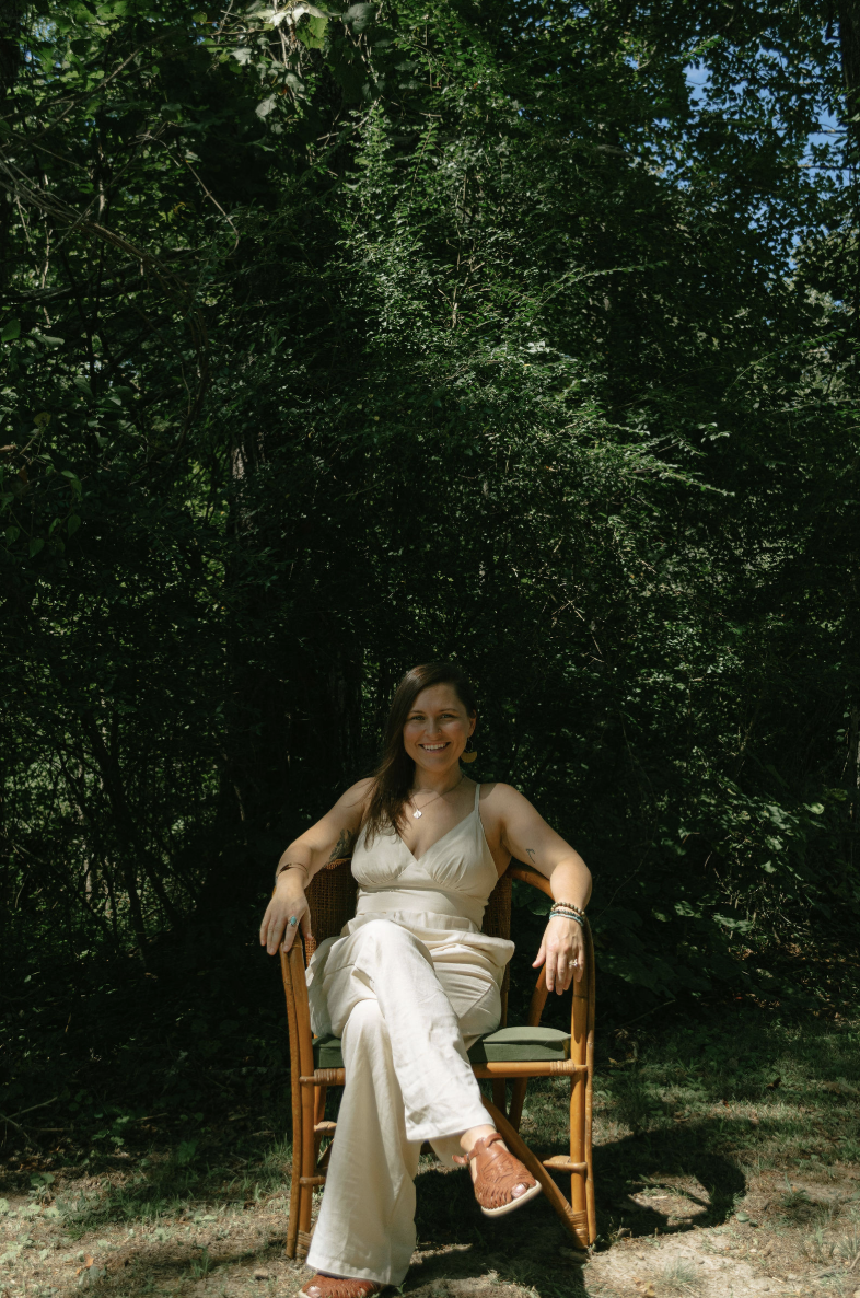 Amberly Kelley-Dotson smiling while seated on a light blue bolster, wearing a light blue button-up shirt tied at the waist over a golden yellow outfit. She sits on a yoga mat in a bright room with wooden floors, with neatly folded blankets beside her